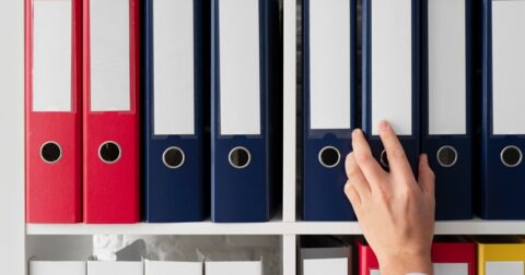 Etagère avec des classeur devant laquelle l'on voit une main qui cherche parmi les classeurs